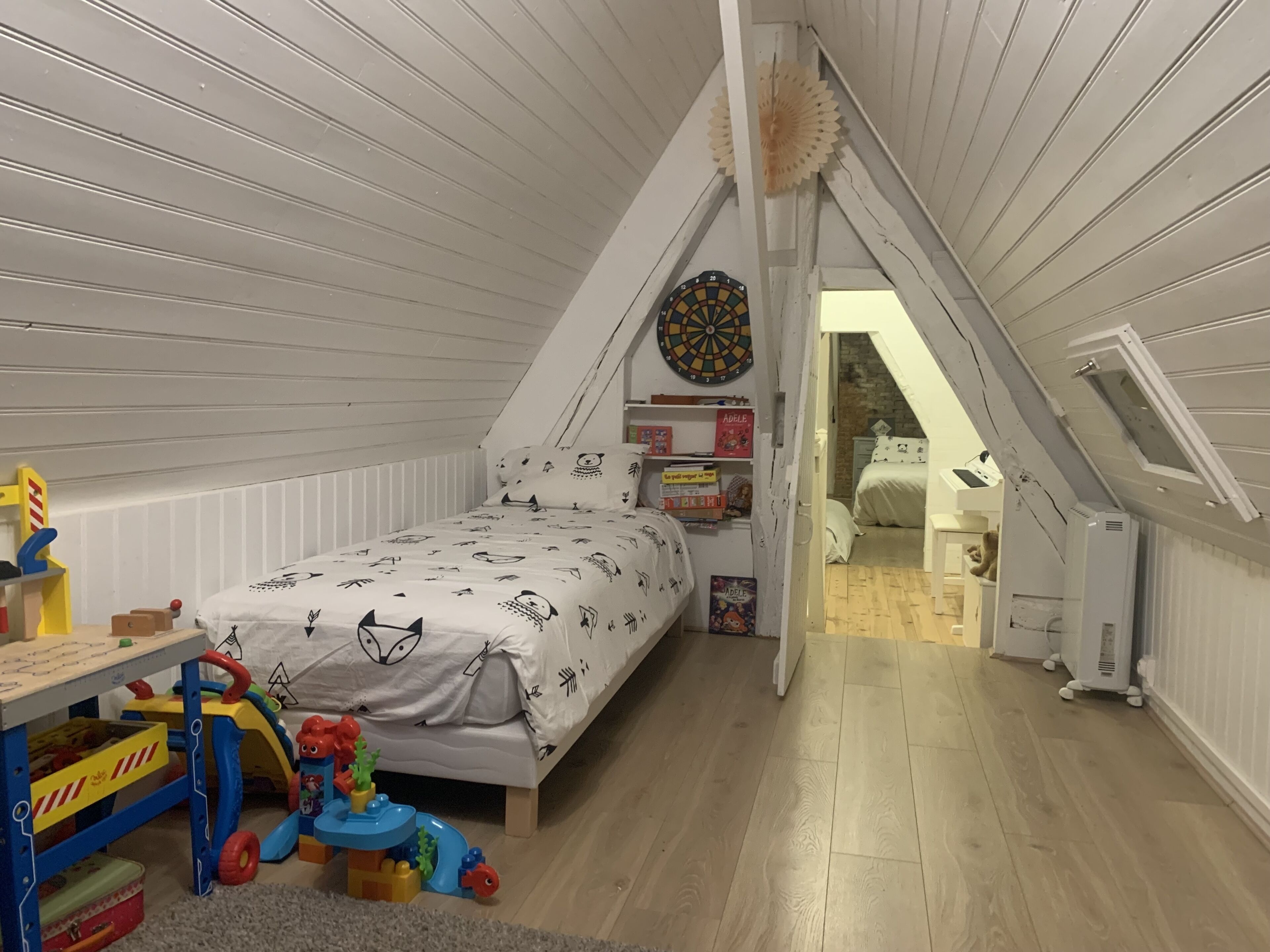 A bedroom with a sloped ceiling, a bed with a white and black animal print duvet, a dartboard, and a bookshelf with children's books.