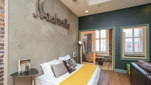A bedroom with a bed, a bedside table, a mirror, and a window with blinds. - 2 Lofts on Broadway Strip - Sleeps 10 (Nashville)