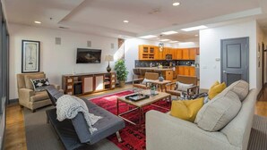 A modern living room with a sofa, armchair, and coffee table. A kitchen with wooden cabinets is visible in the background. - 2 Lofts on Broadway Strip - Sleeps 10 (Nashville)