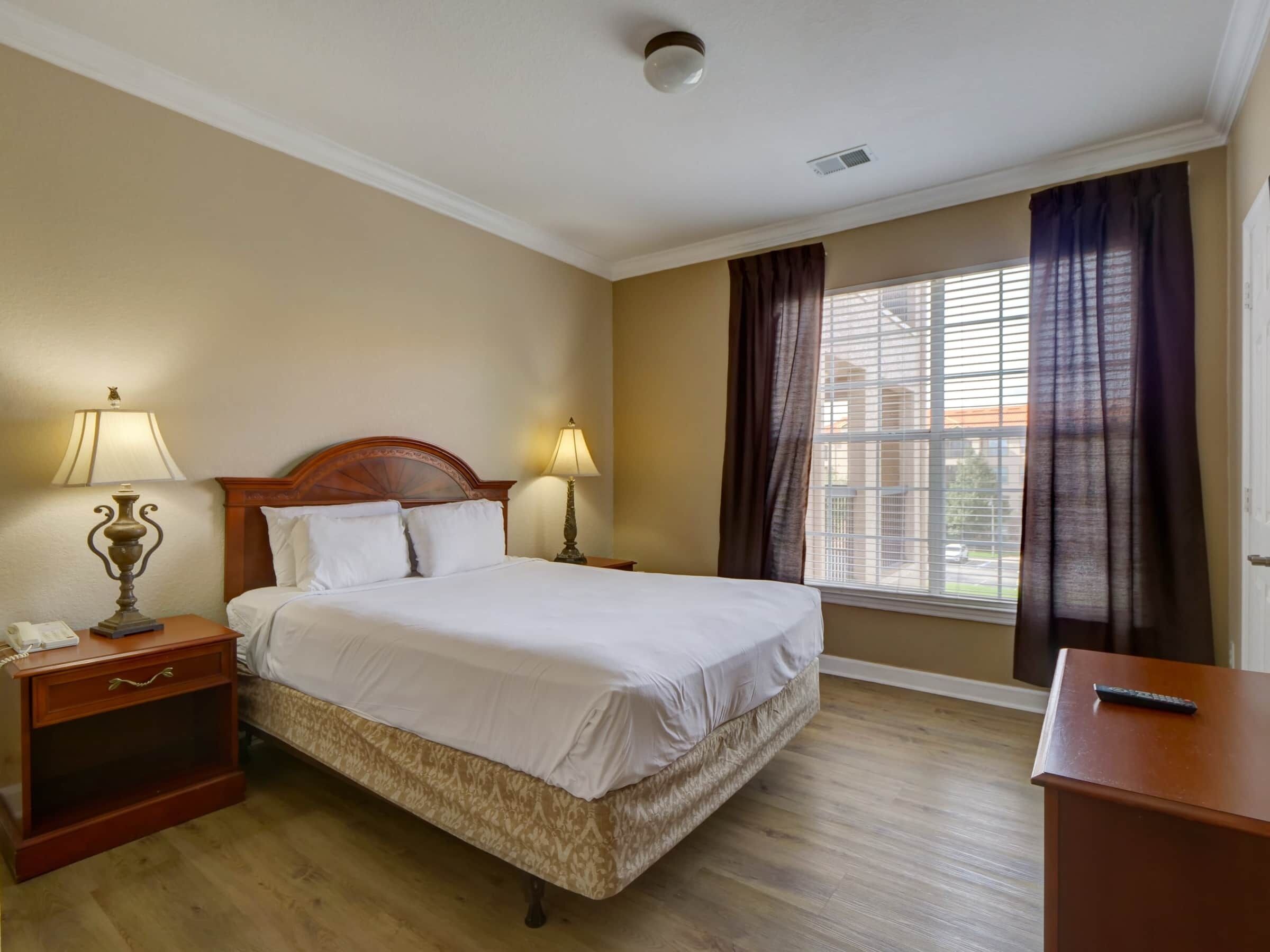 Una habitación de hotel con una cama grande, dos mesitas de noche con lámparas, un tocador de madera y una ventana con cortinas.