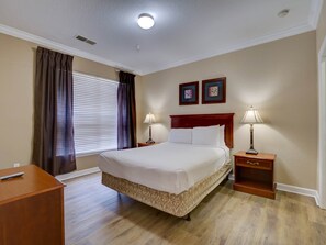 Ein Hotelzimmer mit einem großen Bett, zwei Nachttischen mit Lampen, einer hölzernen Kommode, einem Fenster mit Jalousien und zwei gerahmten Bildern an der Wand.