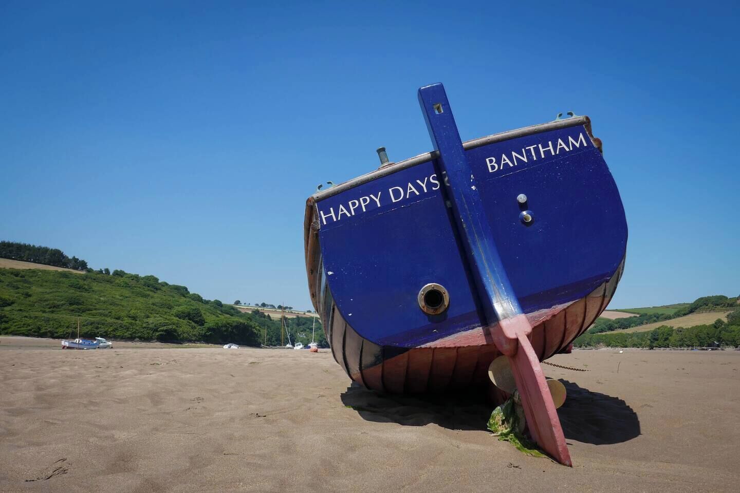 Un bateau bleu sur lequel on peut lire les mots « HAPPY DAYS BANHAM », échoué sur une plage de sable, avec des collines en arrière-plan.