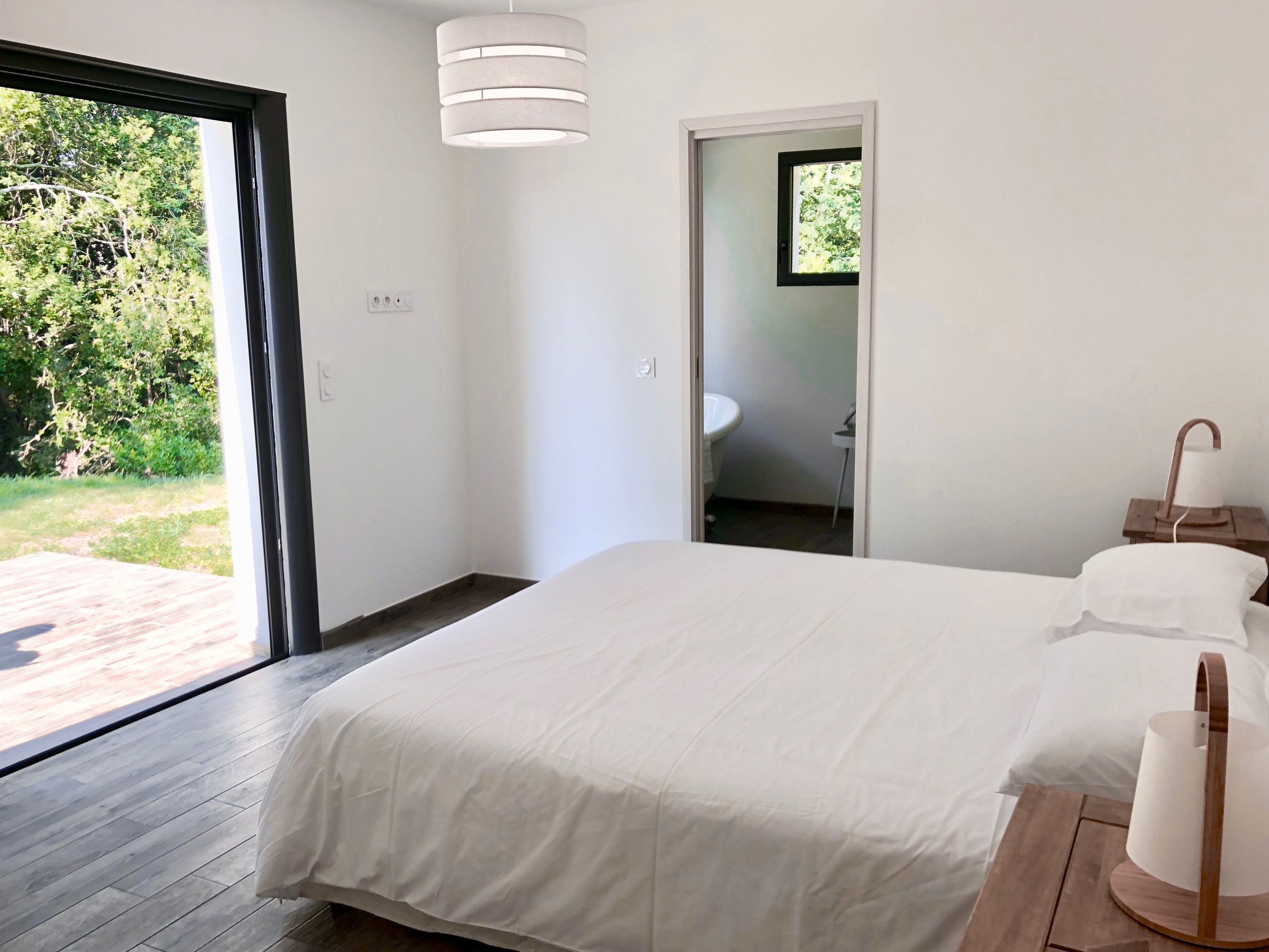 A bedroom with a large bed, a wooden floor, a white ceiling light, and a view of a garden through a glass door.