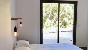 A bedroom with a large bed, a hanging light fixture, and a view of the outdoors through a sliding door. - Villa heated pool & garden •Town center 5 min •Beaches 10 min •Cleaning included (Porto Vecchio)