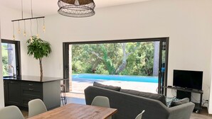 A modern living room with a wooden dining table, grey sofa, and a TV on a stand. A large window offers a view of a swimming pool and greenery. - Villa heated pool & garden •Town center 5 min •Beaches 10 min •Cleaning included (Porto Vecchio)