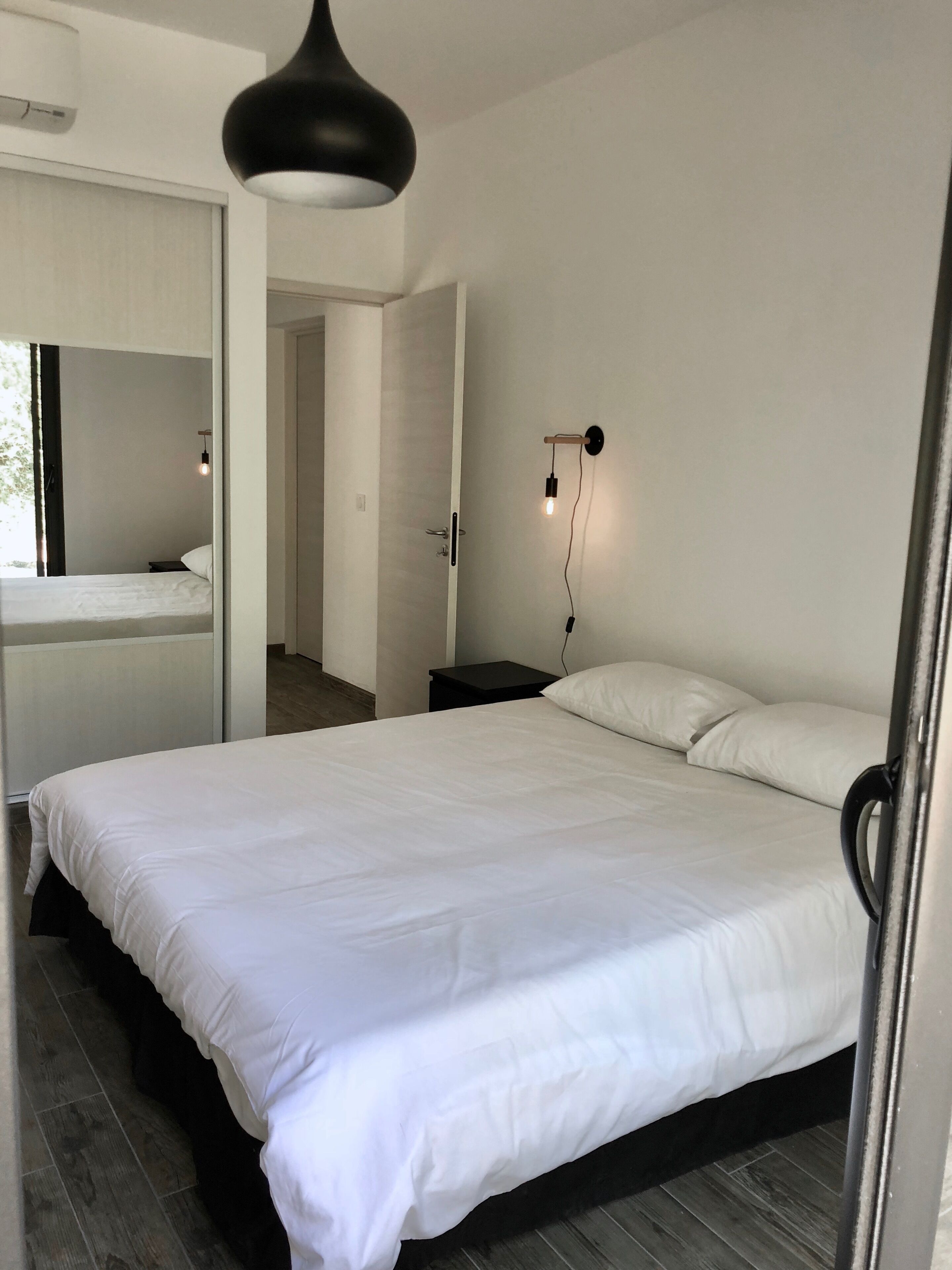 A bedroom with a large bed, a bedside table, a wall-mounted light, and a door leading to another room.