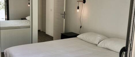 A bedroom with a large bed, a bedside table, a wall-mounted light, and a door leading to another room.