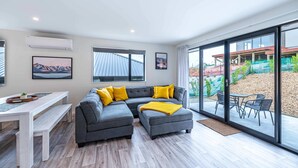 A modern living room with a grey sectional sofa, a white dining table with benches, and a glass door leading to an outdoor patio. - Kirimoko Family Retreat (Wanaka)