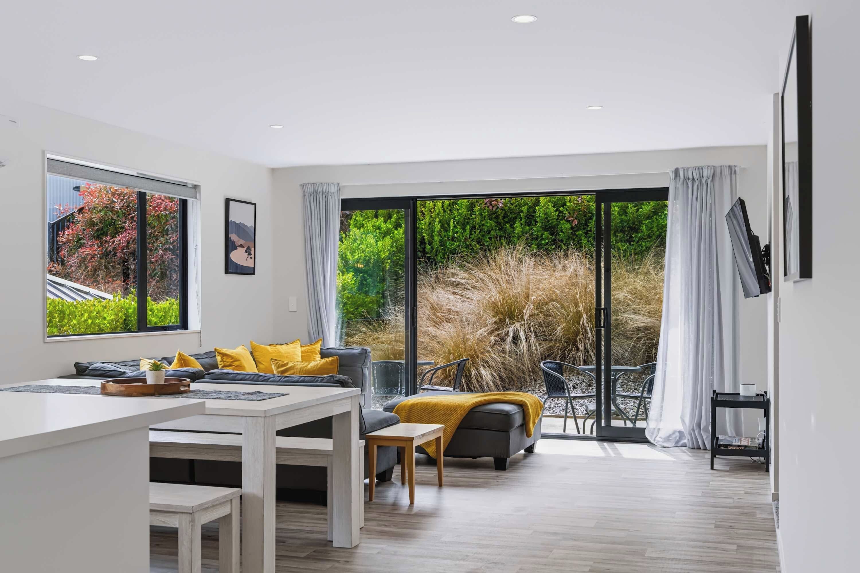 A modern living room with a sofa, dining table, and chairs, large windows, and a view of a garden.