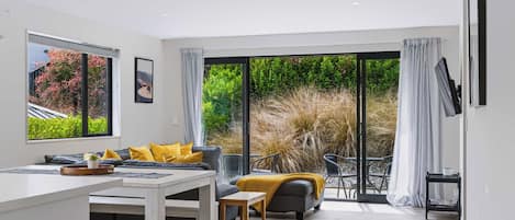 A modern living room with a sofa, dining table, and chairs, large windows, and a view of a garden.