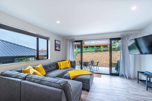 Un salon moderne avec un canapé d’angle gris, des coussins jaunes, un téléviseur à écran plat et une porte-fenêtre coulissante donnant sur une terrasse extérieure.