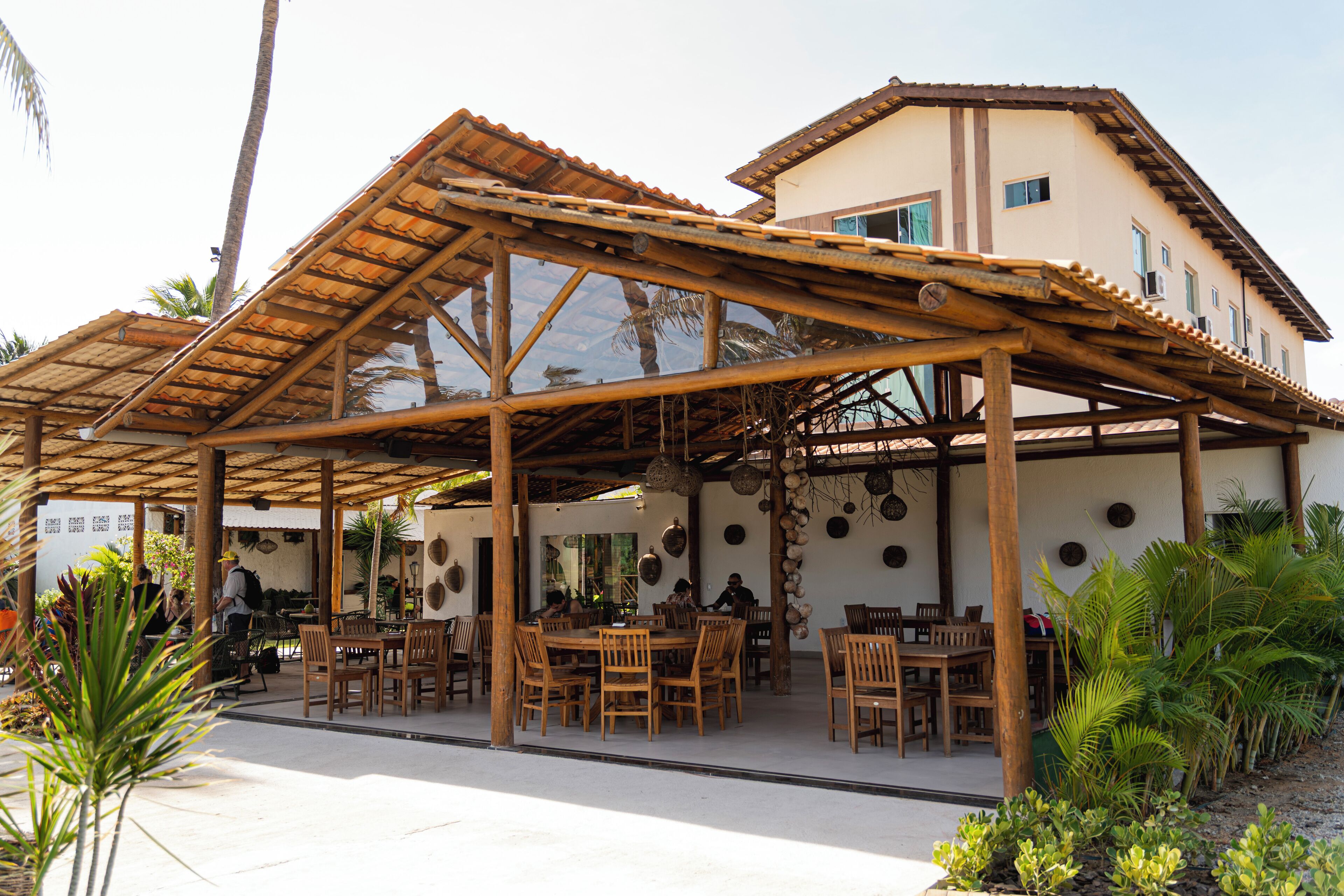 An outdoor dining area with wooden tables and chairs, surrounded by palm trees and a building with a tiled roof.