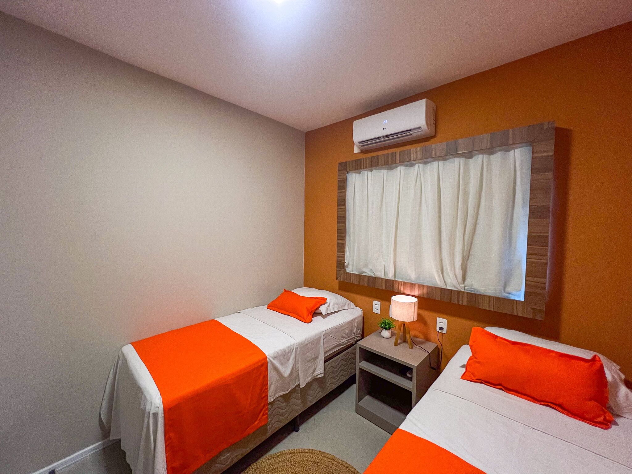Una habitación de hotel con dos camas, cada una con colcha naranja, una lámpara de noche y un aire acondicionado en la pared.