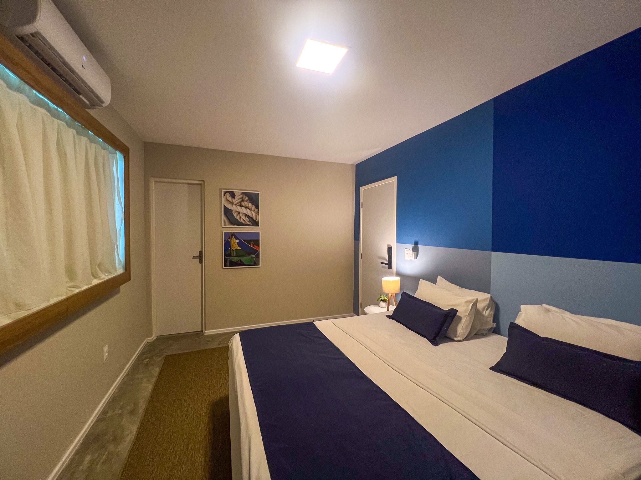 A hotel room with a bed, a bedside table with a lamp, and two paintings on the wall.