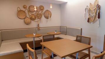 Un interior moderno con una mesa de madera y bancos, una pared decorada con cestas tejidas y joyería de cuentas, y una pequeña planta en maceta sobre una mesa redonda.