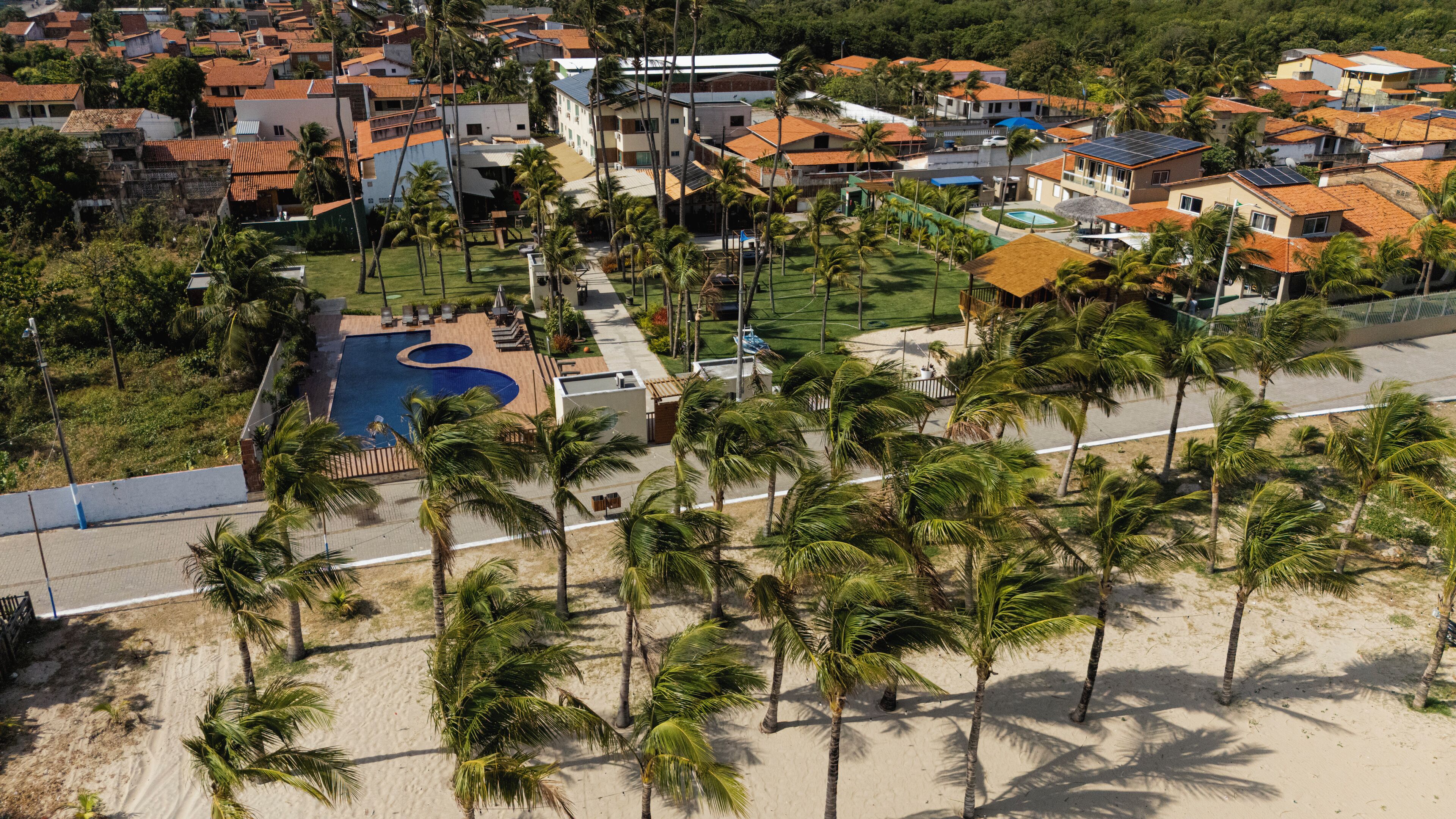 An aerial view of a coastal residential area with houses, a swimming pool, and palm trees.