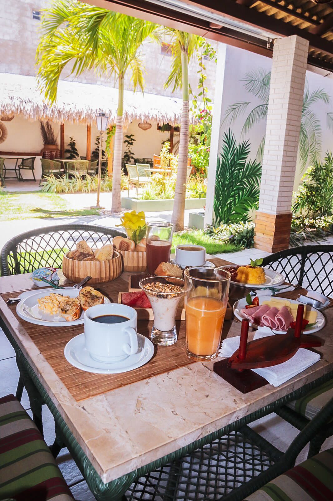 Un desayuno con diversas opciones de comida y bebida sobre una mesa, rodeado de un jardín tropical y una estructura de techo de paja.