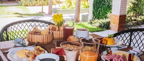A breakfast setup with a variety of foods and drinks on a table, surrounded by a tropical garden and a thatched-roof structure.