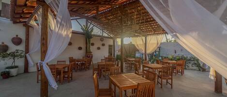 An outdoor dining area with wooden tables and chairs, covered by a wooden pergola with white drapes.
