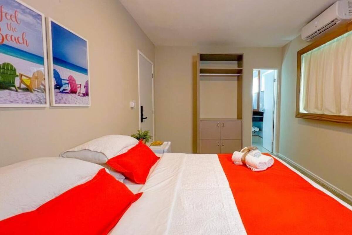 A hotel room with a bed, red pillows, a white bedspread, a red carpet, a mirror, a wardrobe, and a beach-themed painting on the wall.