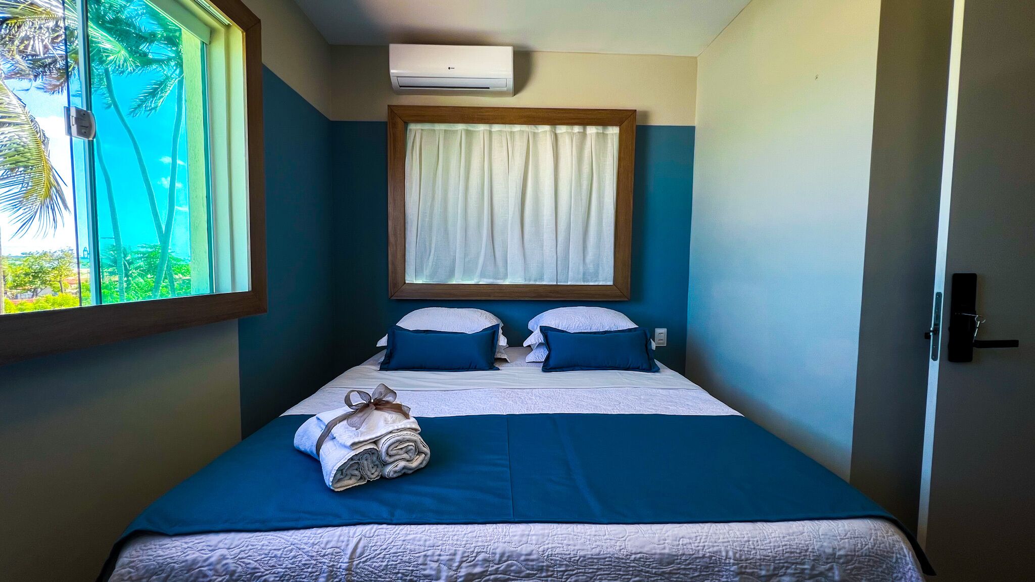 A single bed with a blue and white bedspread, two pillows, a rolled towel, and a window with a view of palm trees.