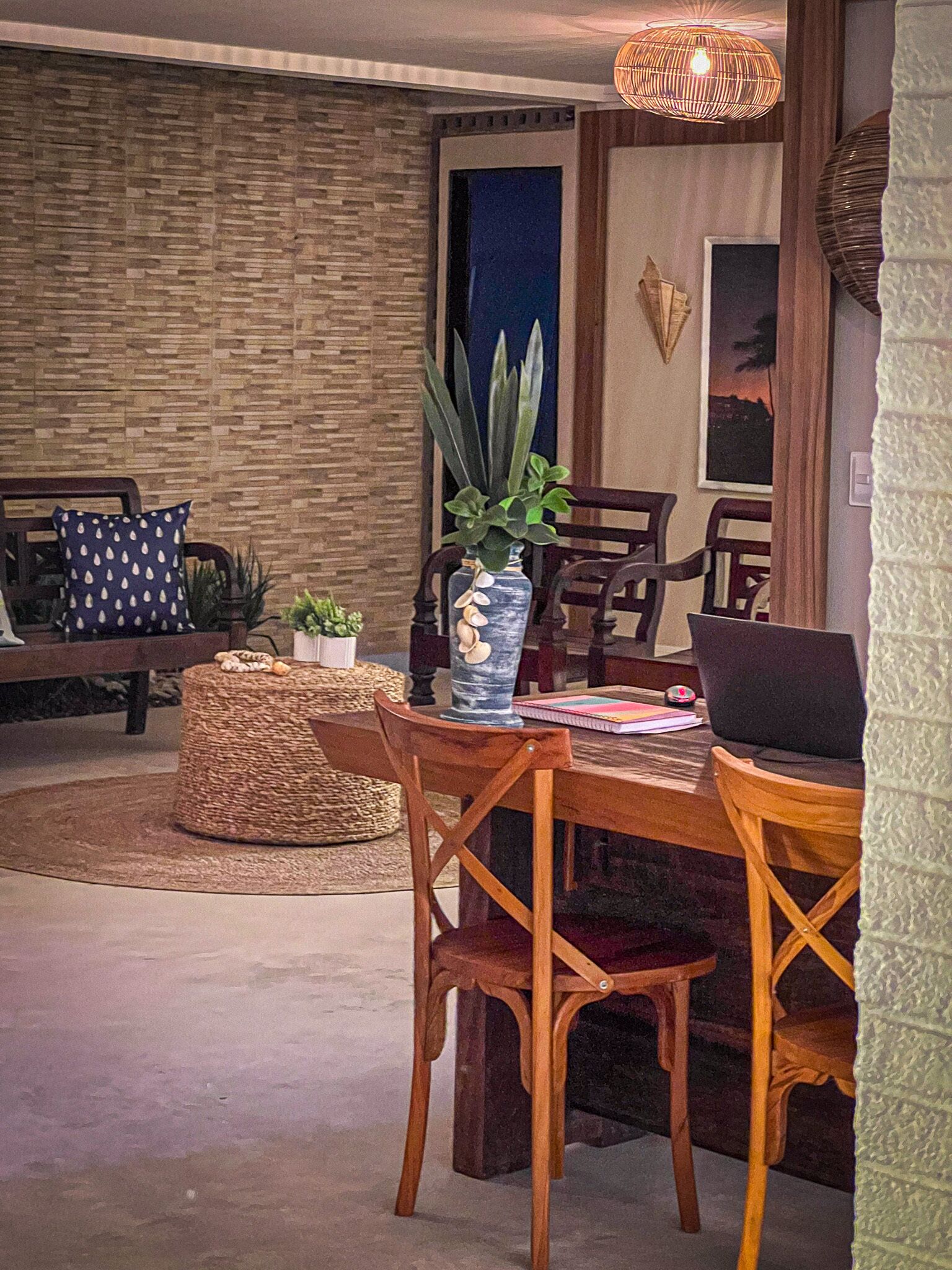 Un área de comedor con una mesa de madera, sillas y un pouf tejido. Hay un jarrón con una planta sobre la mesa.
