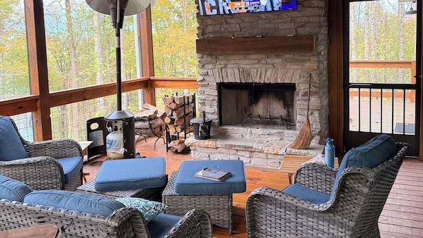 A cozy living room with wicker furniture, a stone fireplace, a television, and a large window overlooking trees.