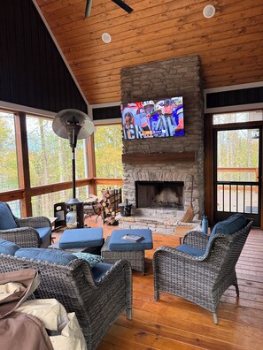 A cozy living room with wicker furniture, a stone fireplace, and a television displaying a sports event. - Nolin Lake/Mammoth Cave Lakehouse Rental - Newly Built (Leitchfield)