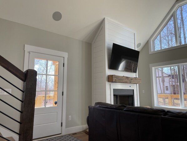 A living room with a fireplace, a TV mounted above it, and a large window providing natural light. - Nolin Lake/Mammoth Cave Lakehouse Rental - Newly Built (Leitchfield)