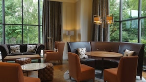 Ein moderner Lounge-Bereich mit Ledersofas, Sesseln und einem runden Tisch, großen Fenstern mit Vorhängen und Blick ins Grüne.