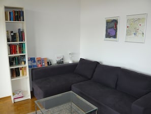 A living room with a dark-colored sofa, a glass coffee table, a white bookshelf filled with books, and two framed abstract paintings on the wall.