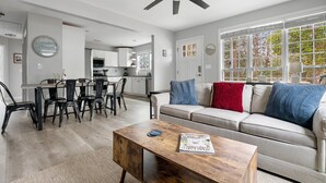A modern living room with a wooden coffee table, a beige sofa with blue and red cushions, a dining area with black chairs, and a kitchen in the background. - Your Dynamic Retreat | Park Circle Paradise (North Charleston)