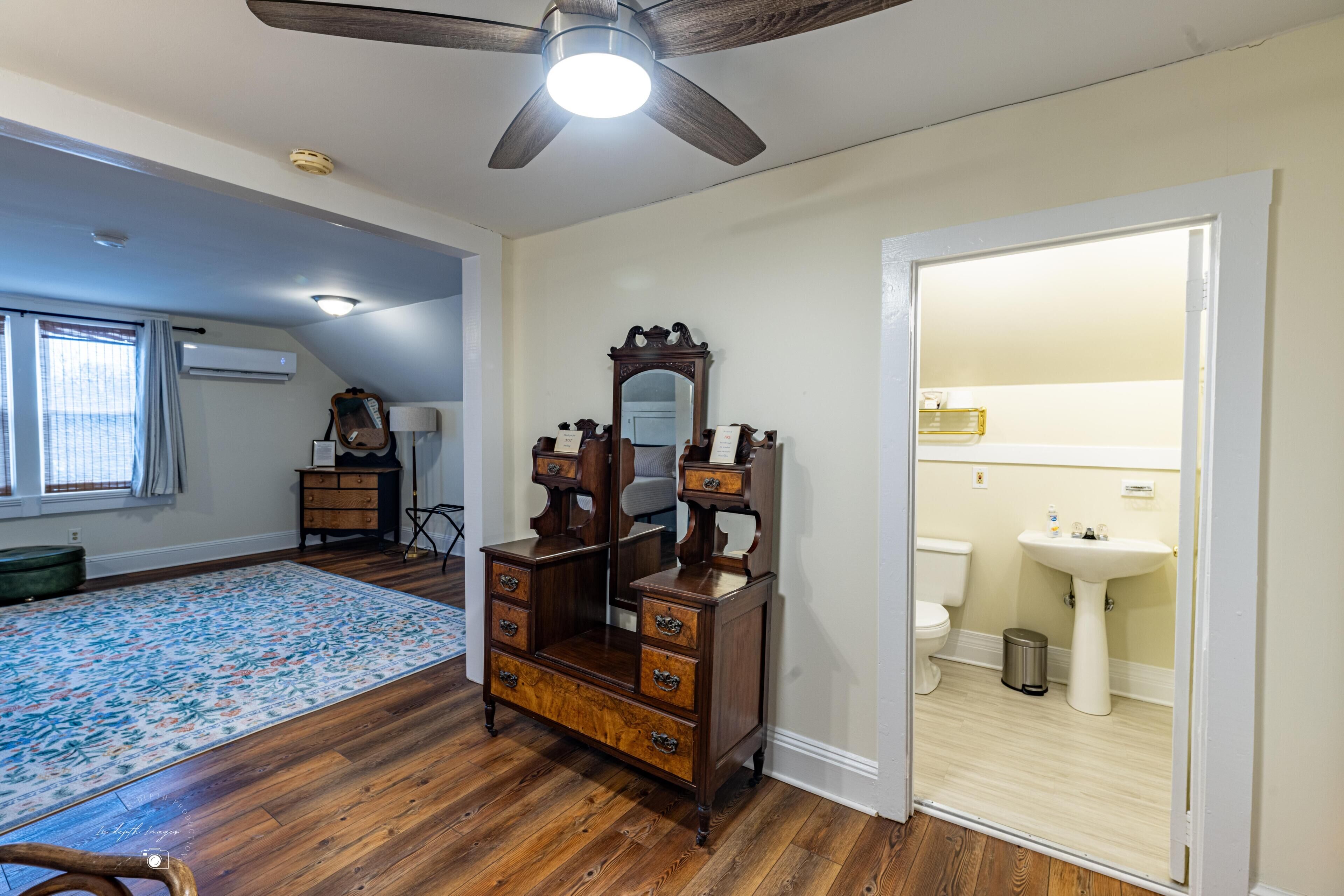 A room with hardwood flooring, a large mirror above a wooden dresser, a ceiling fan, and a window with curtains.