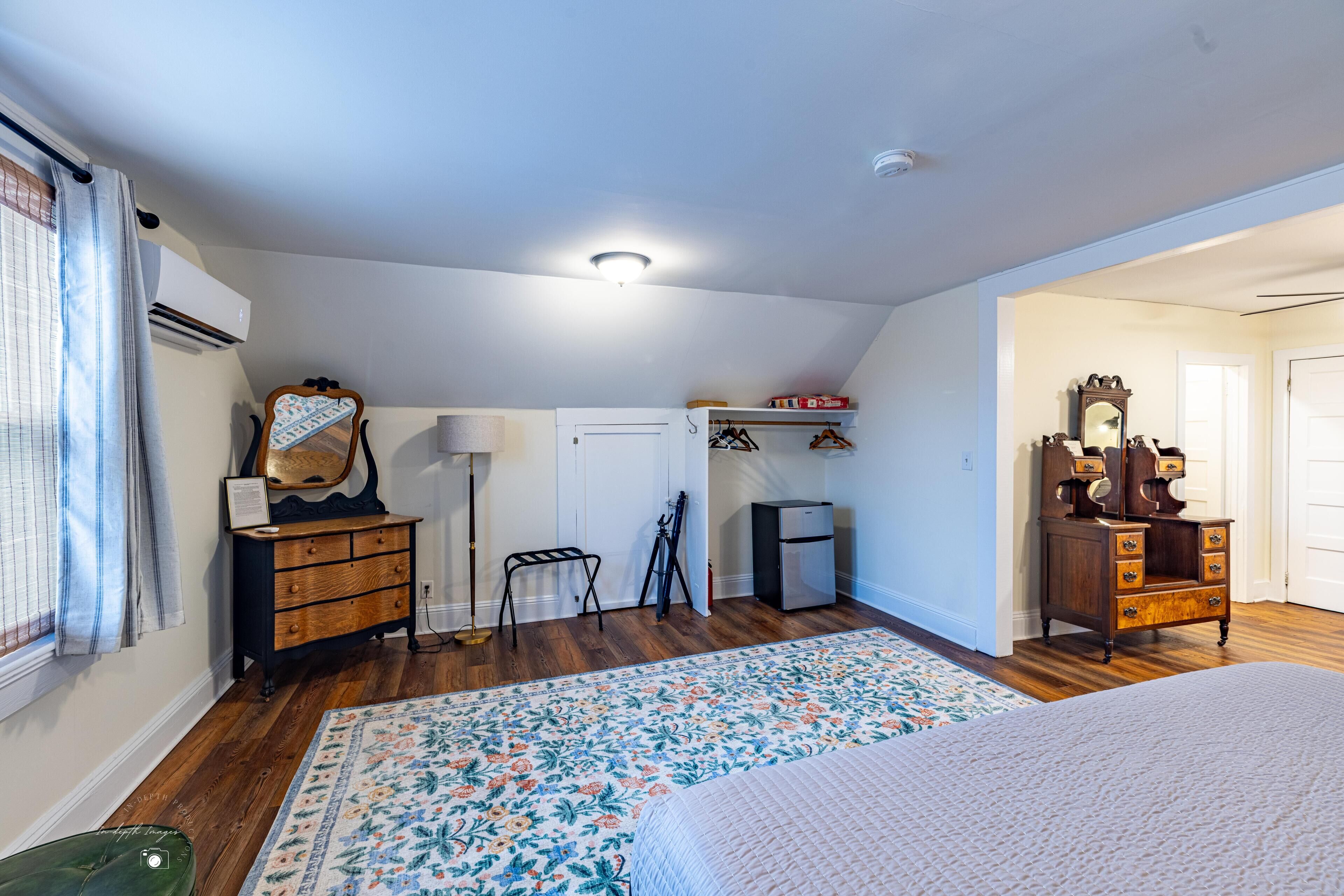 Een slaapkamer met houten vloer, een bed met een bloemenprint, een dressoir, een spiegel en een klein bureau met een lamp.