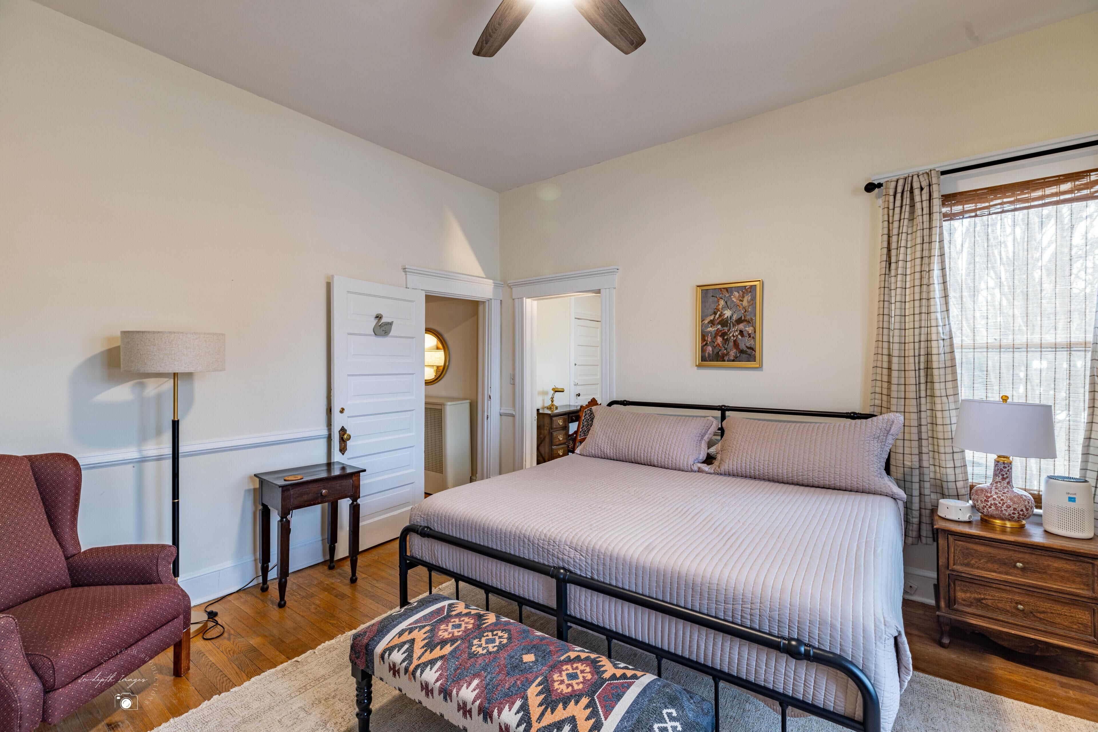A bedroom with a bed, a chair, a bedside table, a lamp, and a window with curtains.