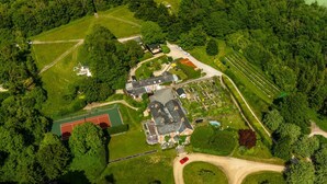 Vue aérienne d’une propriété comprenant une grande maison, un court de tennis et des jardins environnants.