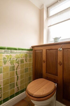 Ein Badezimmer mit einer hölzernen Toilette, Wänden mit grünen und beigen Fliesen und einem Fenster über dem Waschbecken.