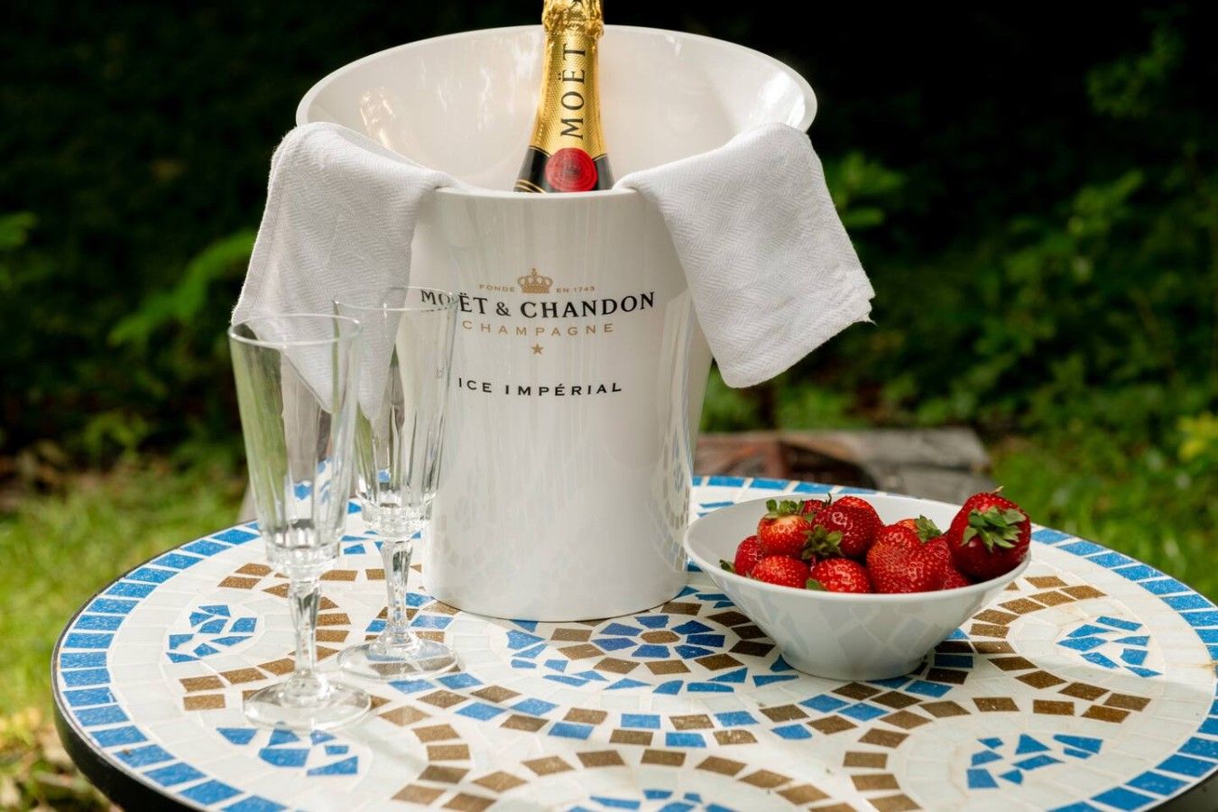 A bottle of Moët & Chandon champagne in a branded ice bucket, a bowl of strawberries, and two glasses on a mosaic table.
