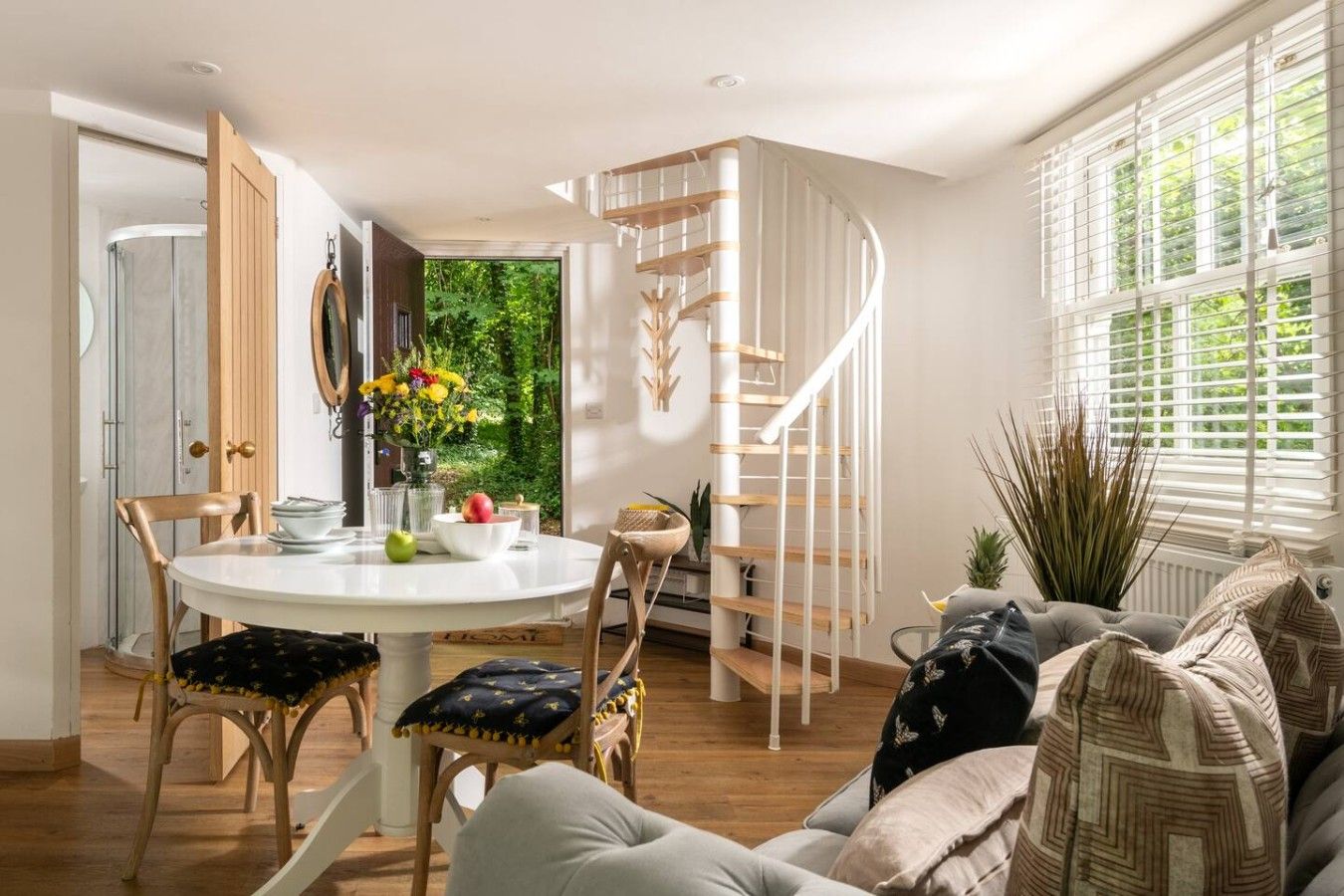 Een modern interieur met een ronde eettafel, houten stoelen, een wenteltrap en uitzicht op groen via een raam.