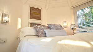 A bedroom with a bed, pillows, a bedside table, a mirror, and a window with blinds. - Honeycomb Treehouse in the Heart of Dorset (Blandford Forum)