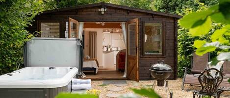 Una cabaña de madera con la puerta abierta que muestra un dormitorio, un jacuzzi y una parrilla.