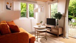 Ein gemütliches Wohnzimmer mit einer Couch, einem Fernseher, einem kleinen Tisch mit einer Schale und einem Fenster mit Blick auf Grünflächen.