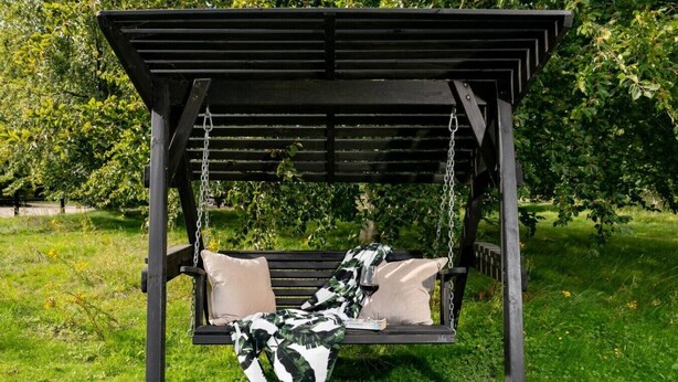 Een houten schommelstoel met kussens, geplaatst in een tuin met gras en bomen.