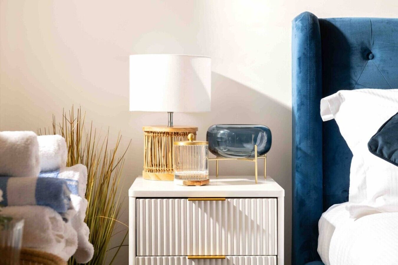 Ett välordnat sovrum med en blå klädd sänggavel, ett sängbord med en lampa, en glasbehållare och en hög med handdukar.