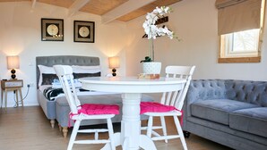 Une pièce chaleureuse avec une table ronde blanche, deux chaises blanches, un canapé gris et un lit avec une tête de lit grise.