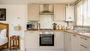 A compact kitchen with a stove, oven, and microwave. There is a small side table with a lamp and a trash bin.
