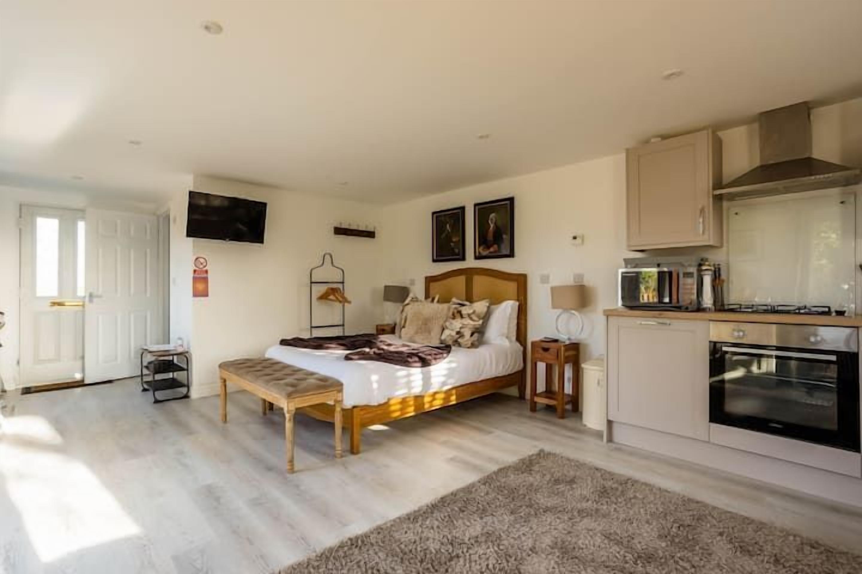 Une chambre à coucher comprenant un lit, des tables de chevet, un banc, une télévision et une kitchenette équipée d’un four.