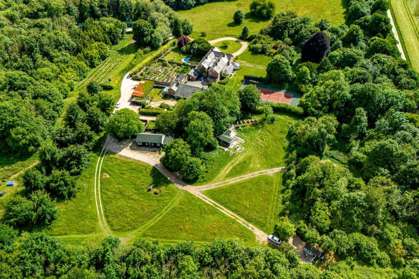An aerial view of a property with multiple buildings, a tennis court, and surrounding green fields.