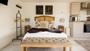 A bedroom with a large bed, bedside tables, a wooden bench, a TV, a coat rack, and a kitchen area.