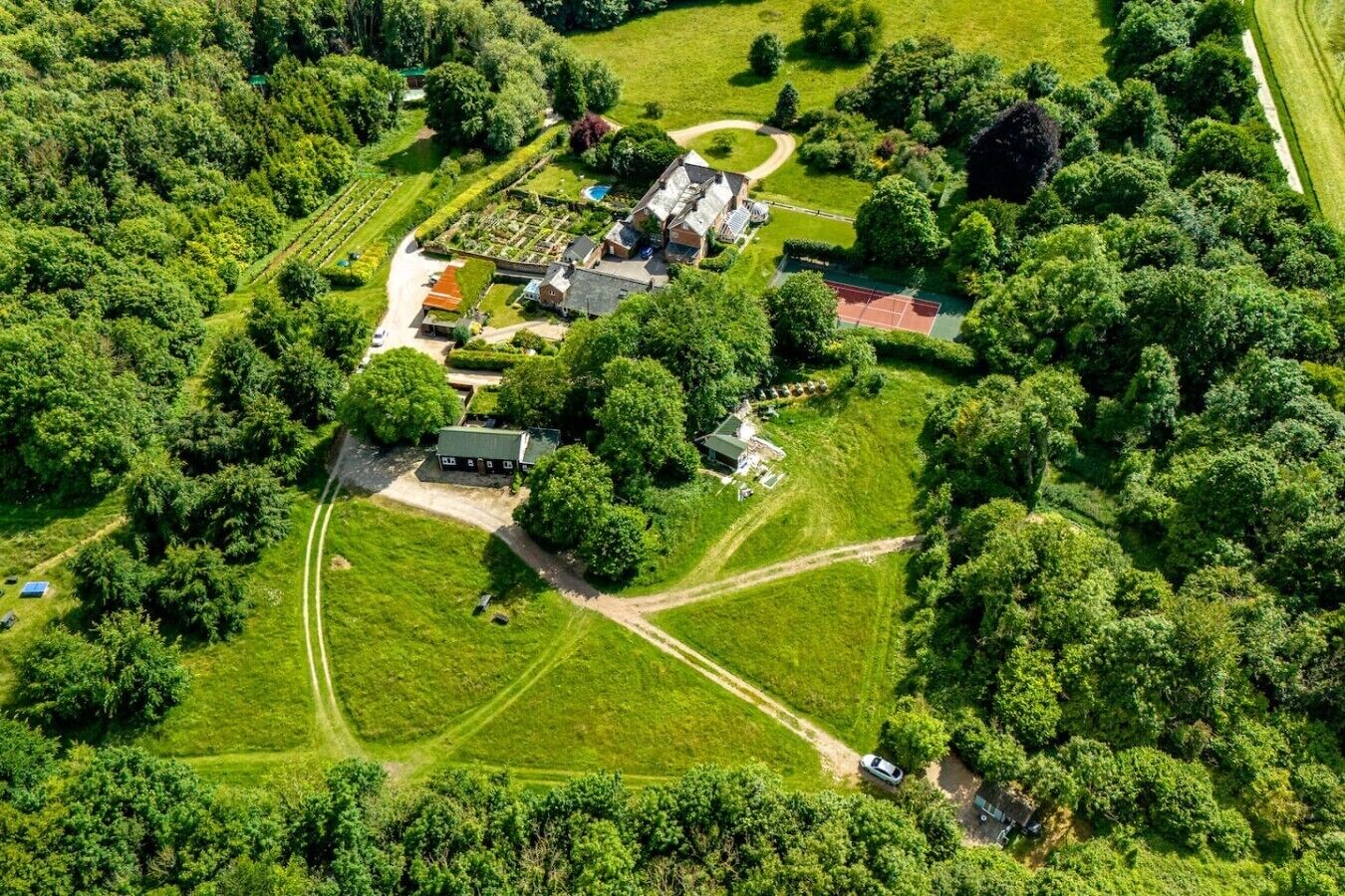 An aerial view of a property with multiple buildings, a tennis court, and surrounding greenery.